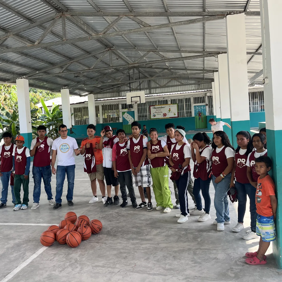 Group of students and adults standing in a semicircle on a covered basketball court, with basketballs in the center and most people wearing matching maroon jerseys labeled "RR".