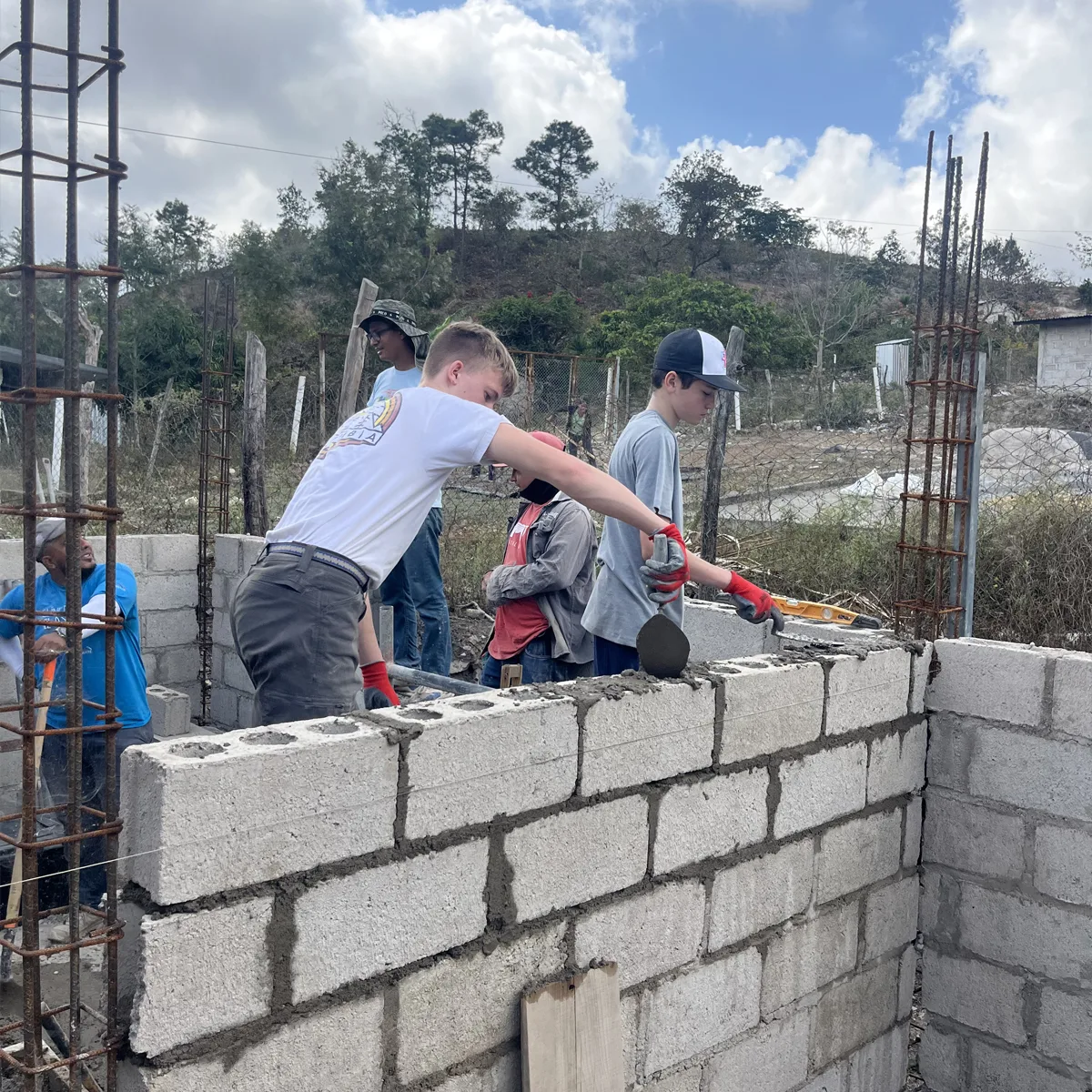 Teen volunteers laying concrete blocks and applying mortar while helping build a cinder block wall at a construction site in a rural area, with others assisting in the background.