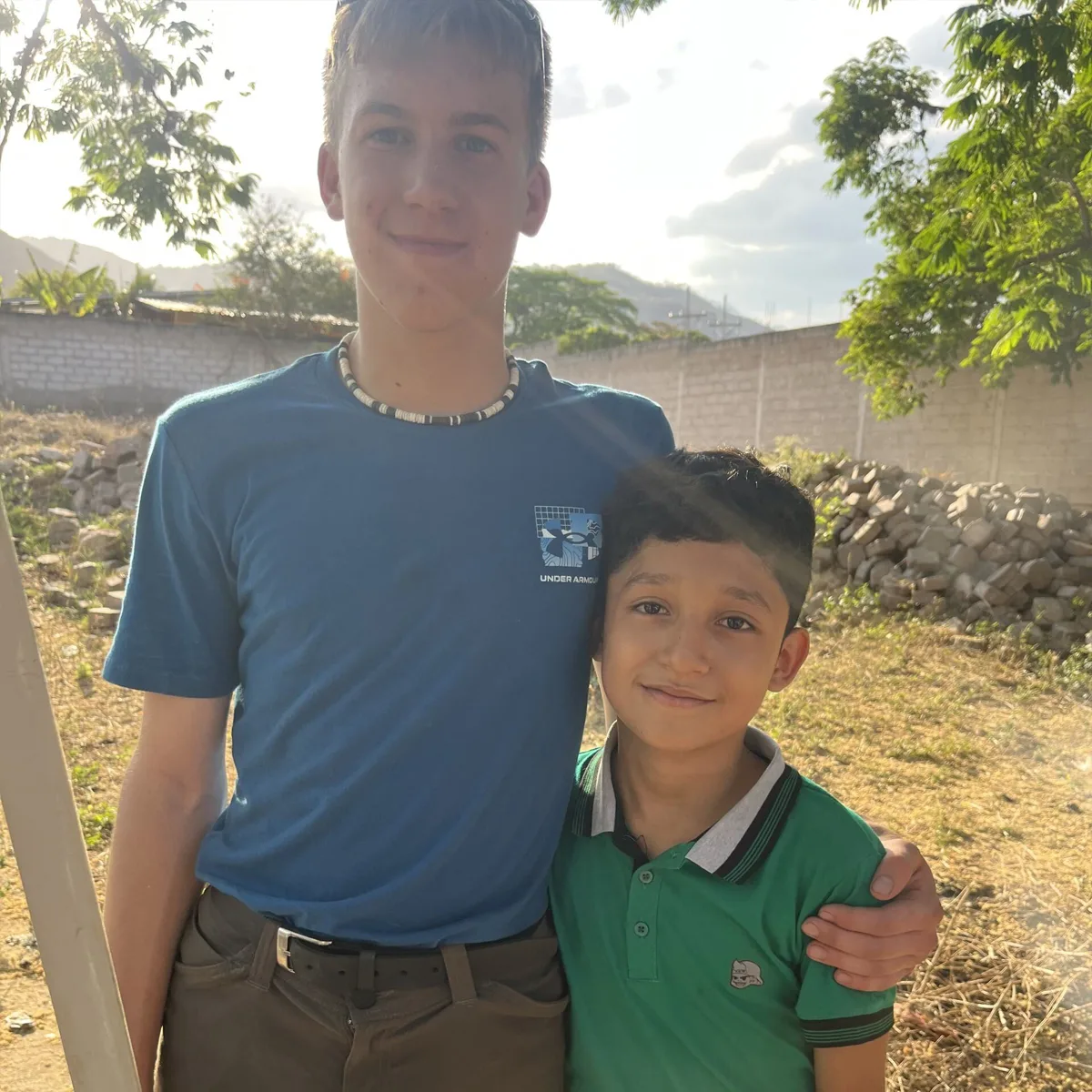 Two boys smiling and posing outdoors in sunny weather; the older wears a blue Under Armour t-shirt, the younger a green collared shirt. They stand near a stone pile with trees and mountains in the background.