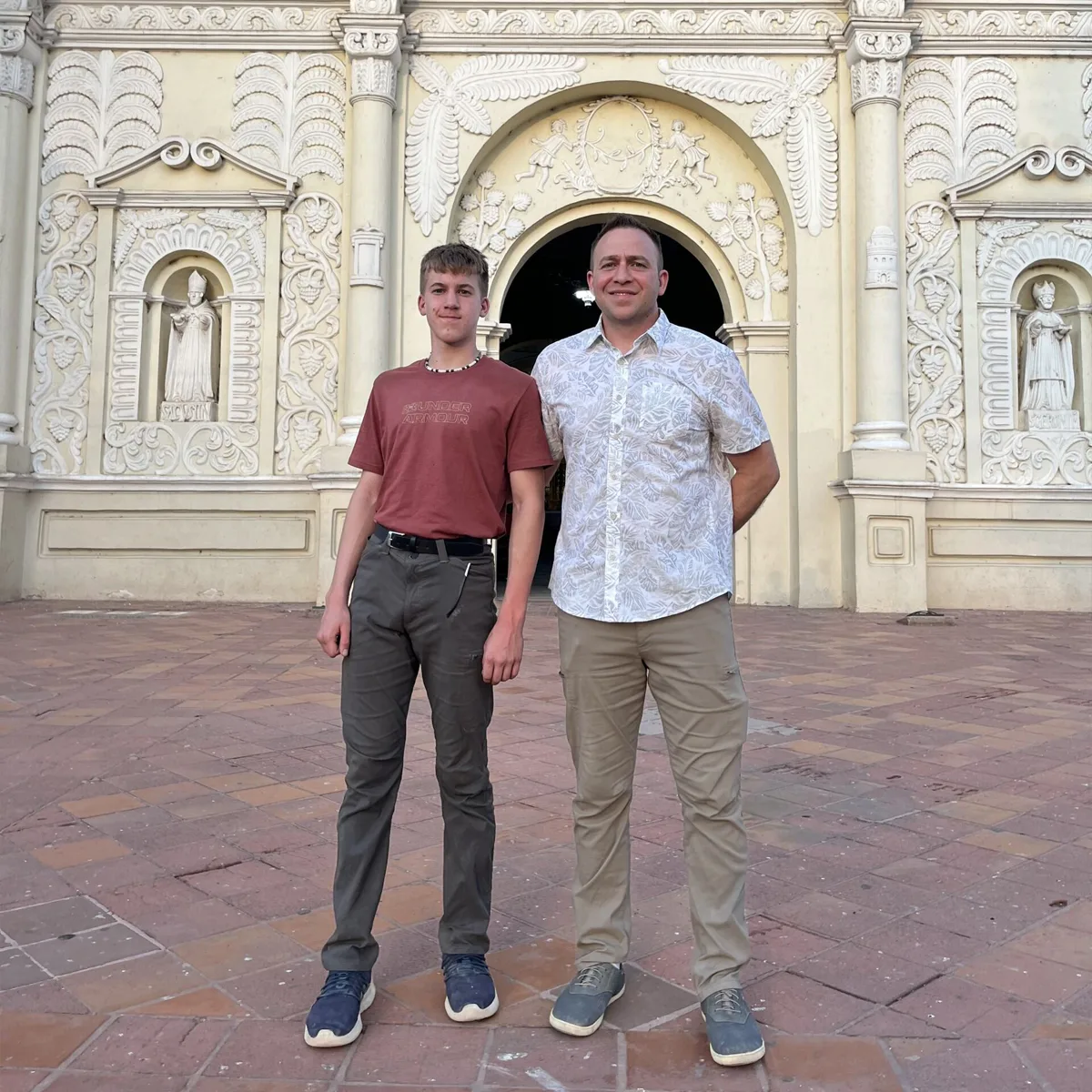 Two men standing and smiling in front of an ornate historic building with carved statues and decorative stonework; one wears a red t-shirt and the other a white patterned shirt.