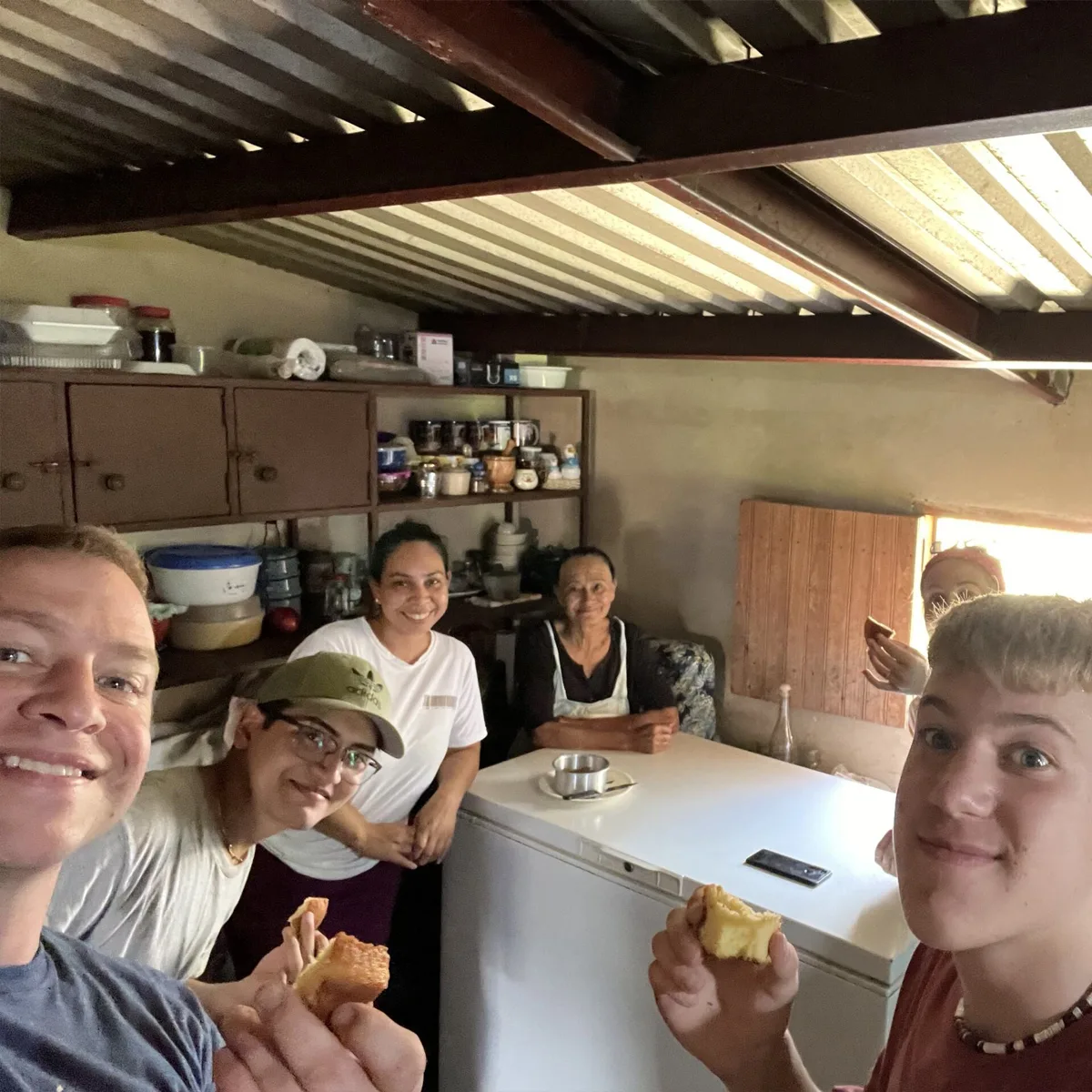 Group of six people smiling inside a modest kitchen, some holding pieces of cake; shelves with jars and containers line the wall behind them, and a chest freezer stands in the center.