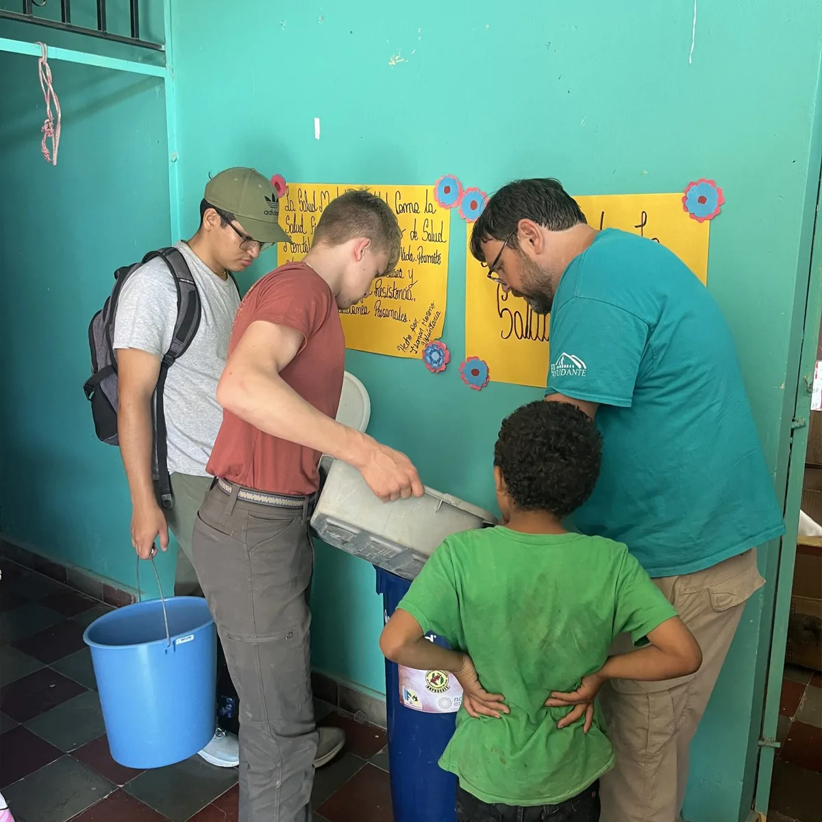 Four people, including a child, working together to pour water into a blue container indoors; one person holds a blue bucket while colorful posters with Spanish text decorate the teal wall behind them.