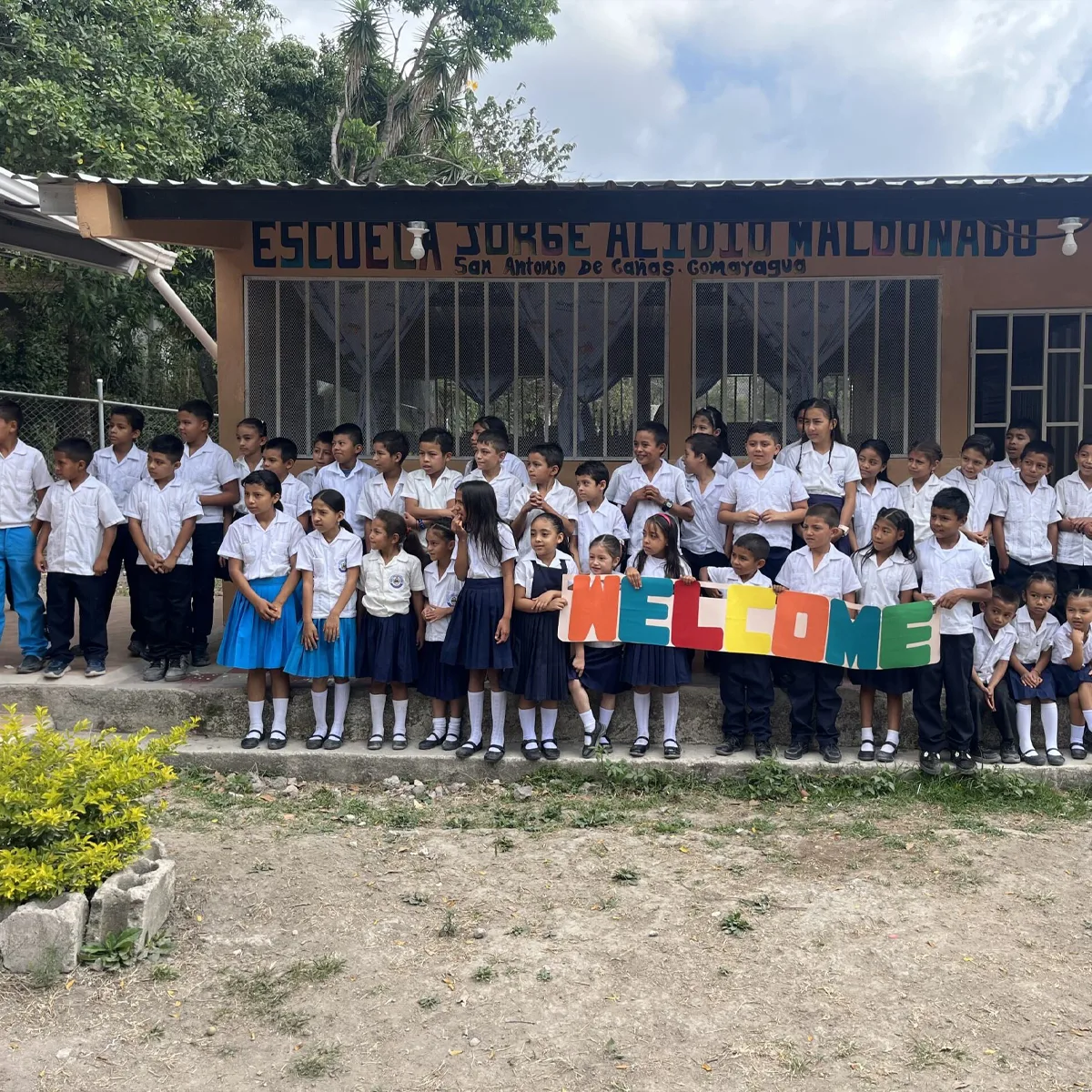 Group of schoolchildren in uniform standing in front of Escuela Jorge Alidio Maldonado, holding colorful letters spelling “WELCOME” with trees and a school building in the background.