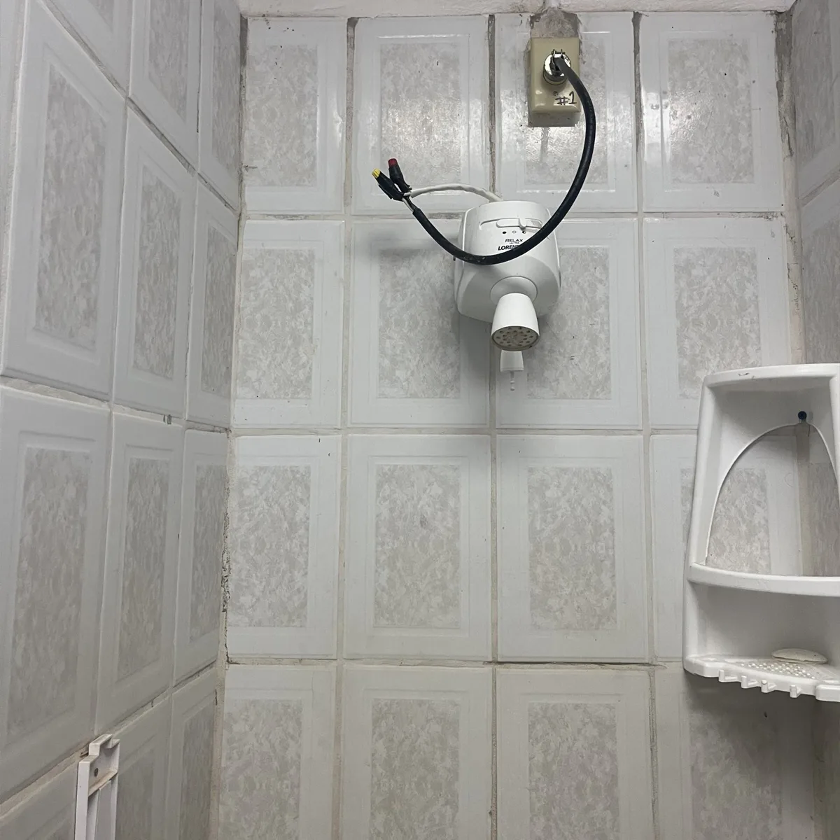 lose-up of a tiled shower with a basic electric showerhead connected to exposed wires, mounted near a white plastic corner shelf holding a bar of soap.