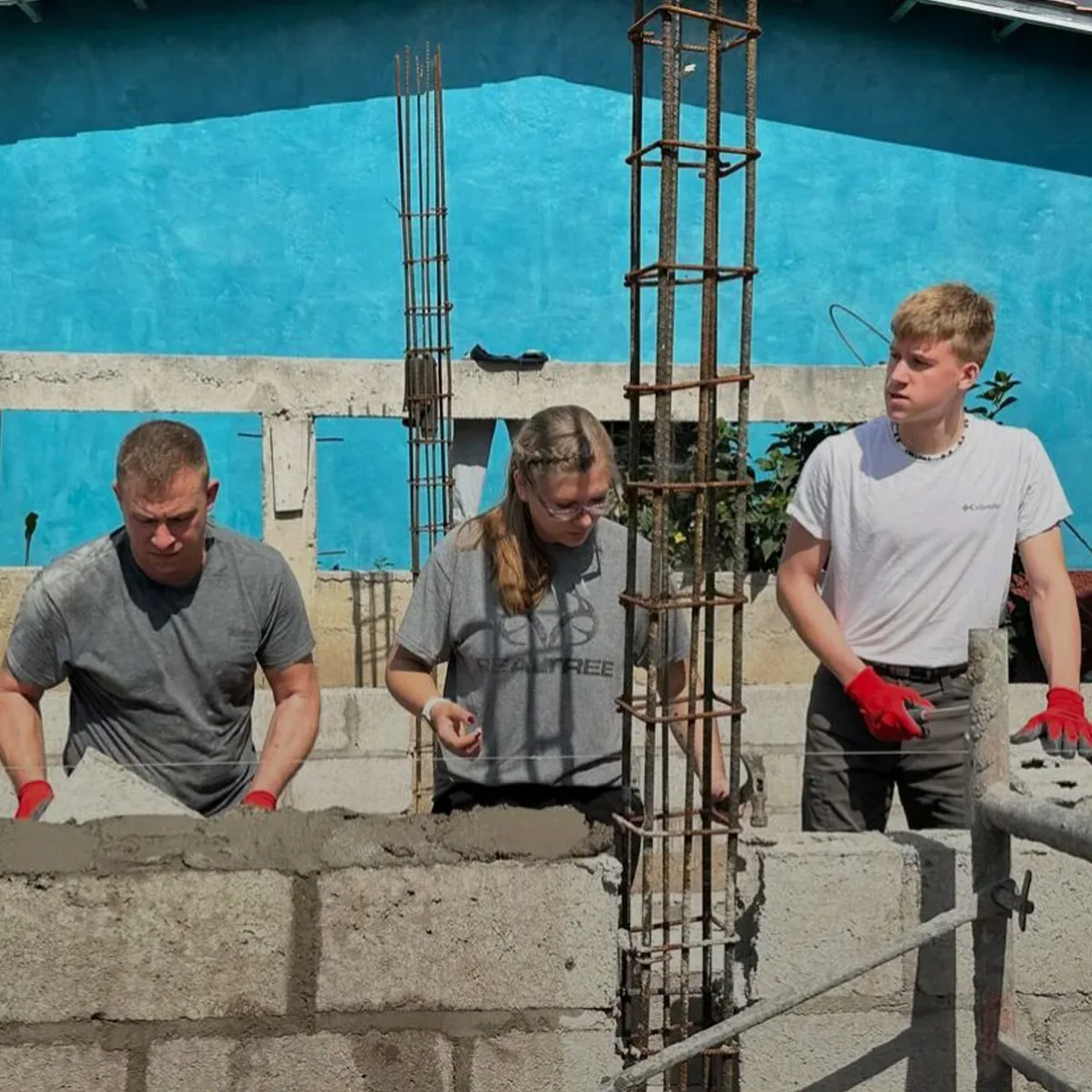 Three people working on a concrete block wall under a bright blue sky. Two young adults and an older man are using trowels to apply mortar between blocks. Rebar columns are rising from the unfinished wall, and all are wearing work gloves and casual clothes.