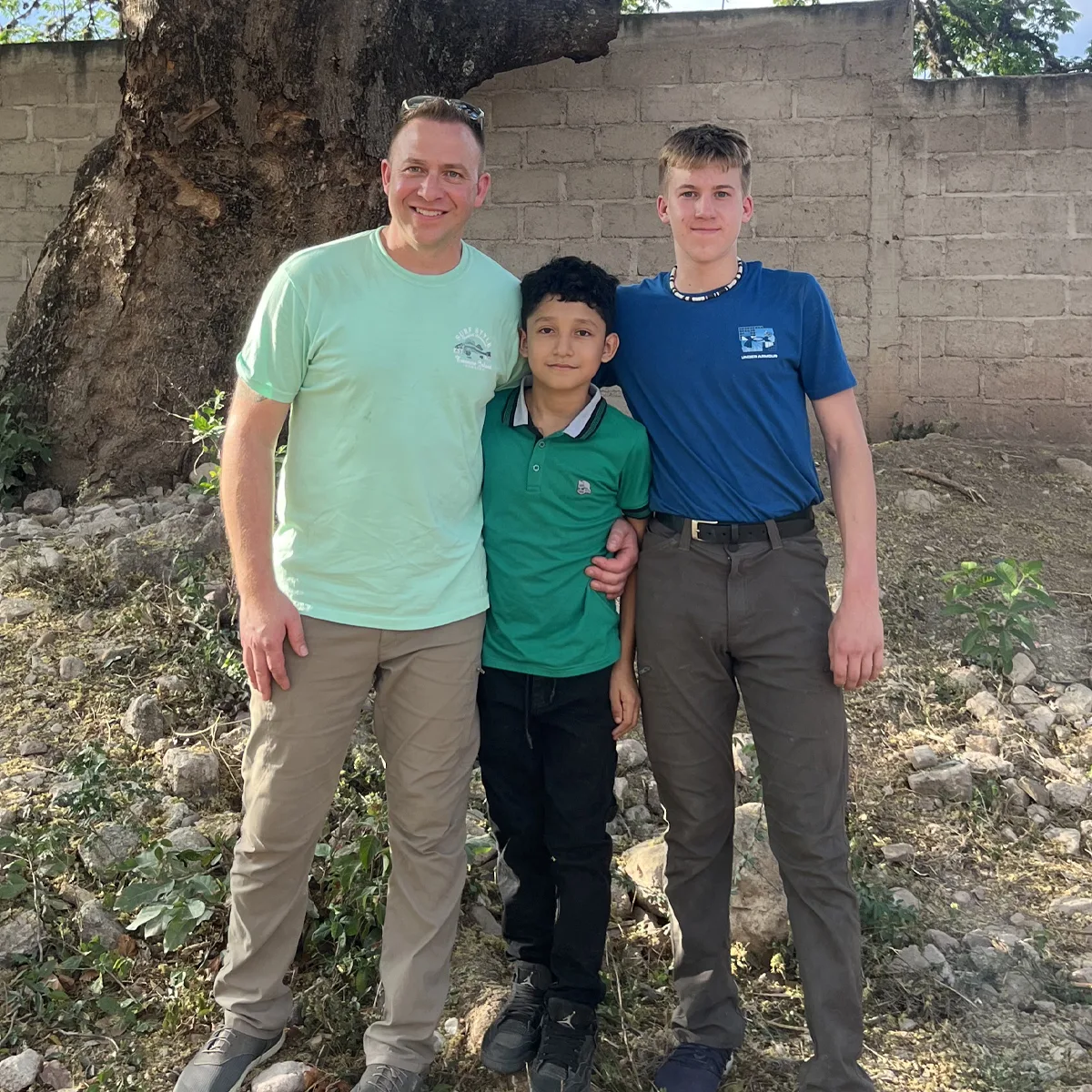 Two mission trip volunteers standing with a local boy in front of a large tree and brick wall, smiling for the camera.