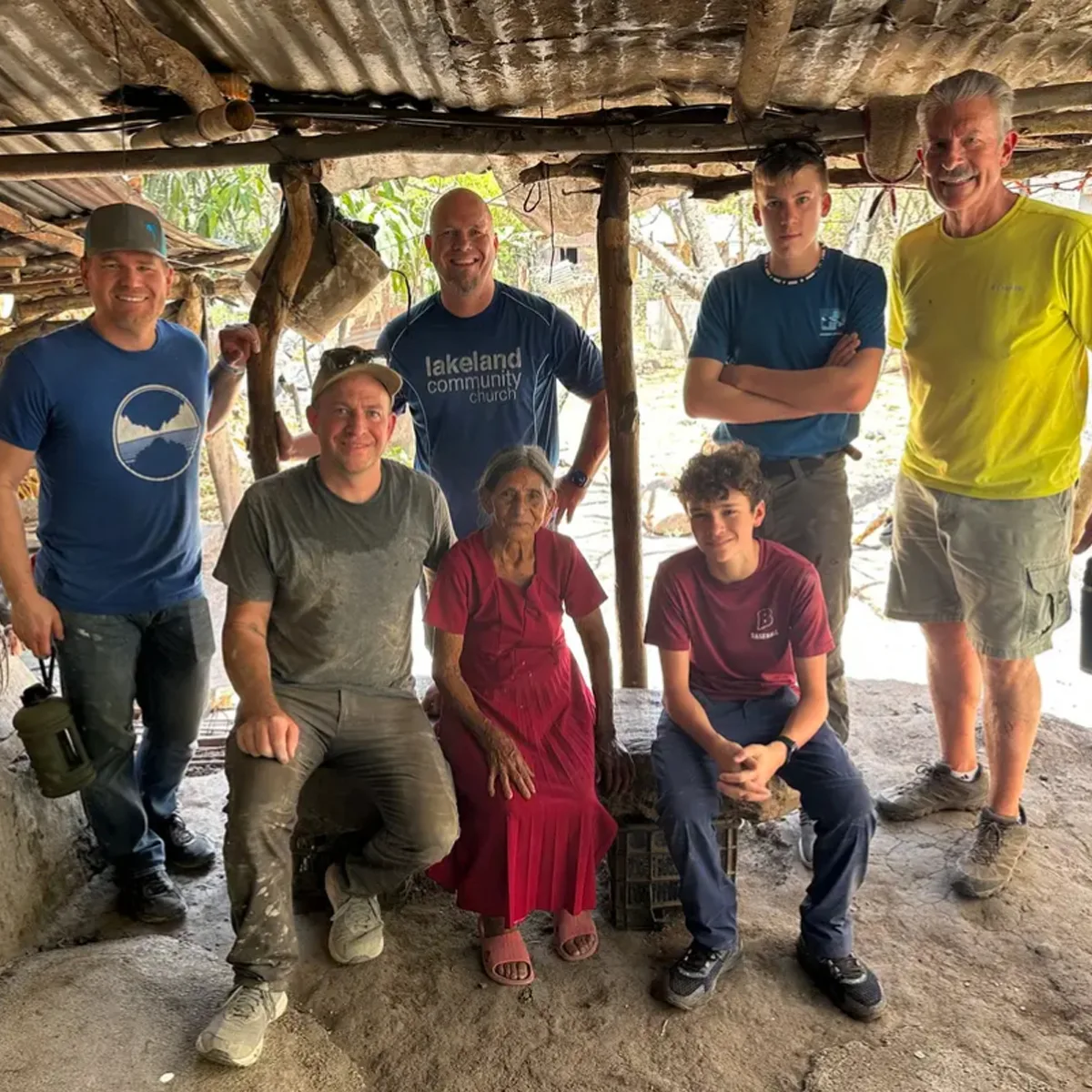 Group of mission team members posing with an elderly woman inside a rustic, open-air shelter with a thatched roof.