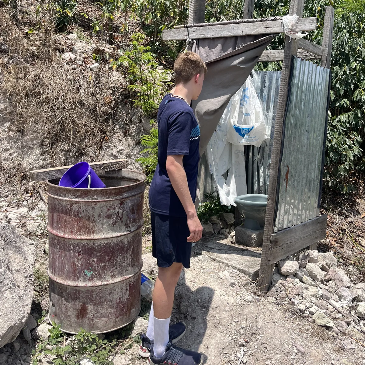 Teenage boy in navy blue shirt and shorts standing near a makeshift outdoor toilet with a tin sheet wall and plastic curtain, next to a rusty barrel with purple buckets.