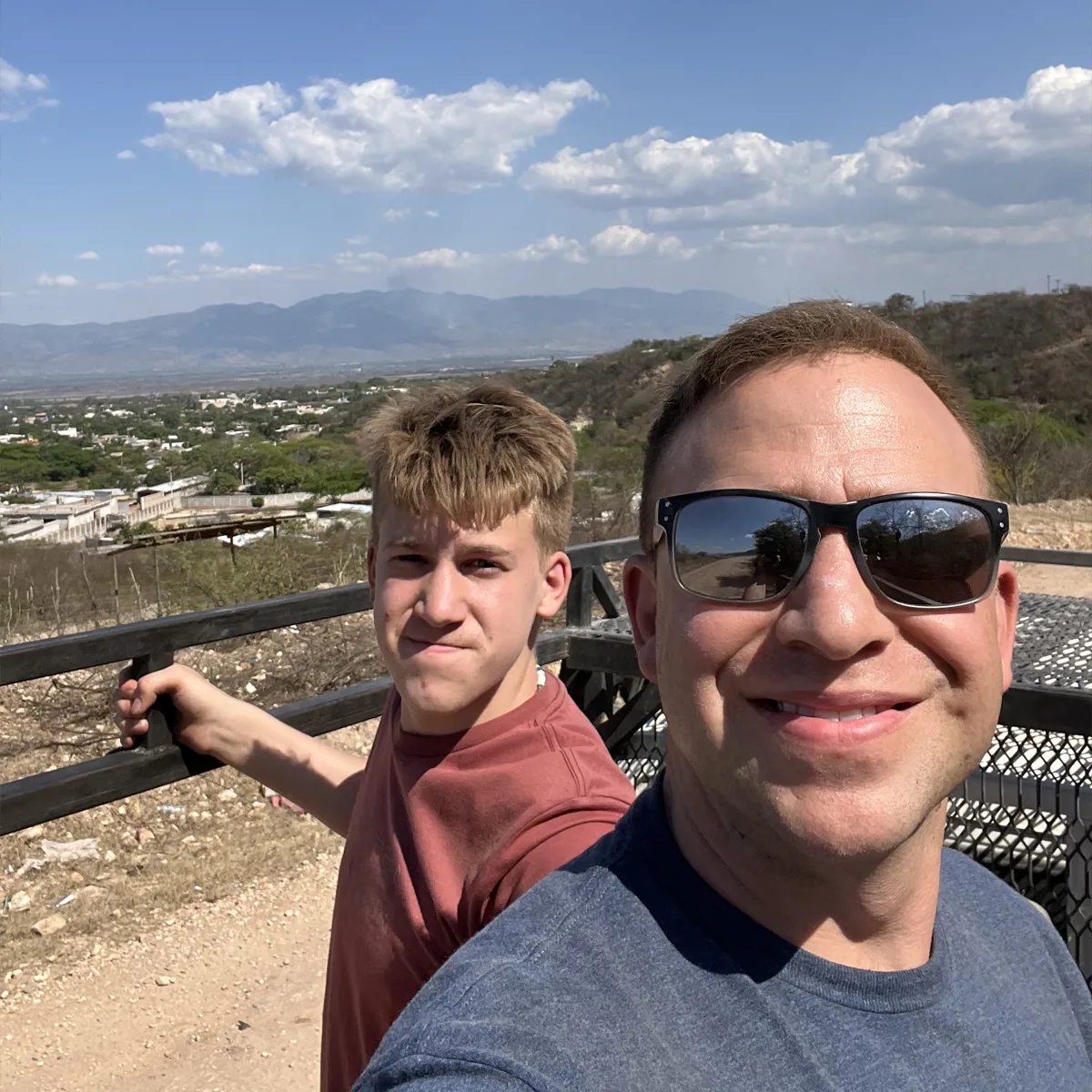 Man in sunglasses and a teen boy take a selfie outdoors with a scenic mountain and valley landscape in the background under a bright blue sky with clouds.
