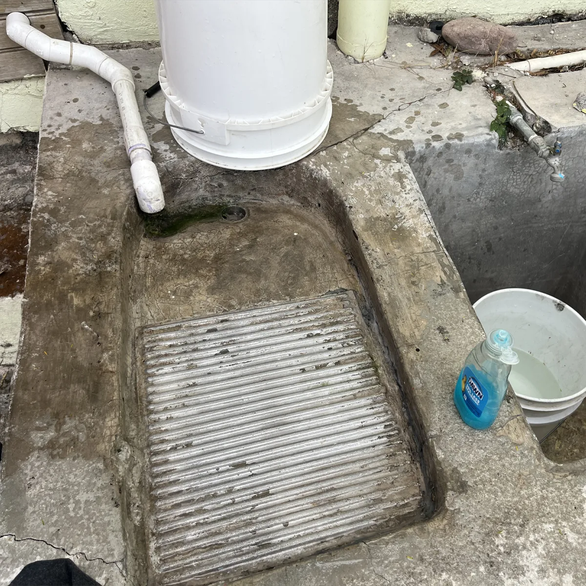 Concrete outdoor wash station with a ridged scrubbing surface, a bottle of dish soap, white plastic bucket with water, and visible PVC piping.